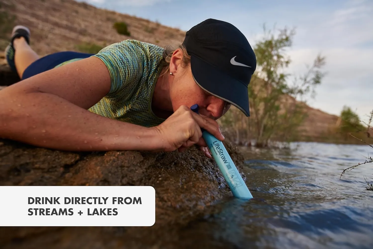 Woman using LifeStraw personal water filter to drink directly from a stream during a hike, demonstrating portable water purification for outdoor survival, camping, and emergency preparedness.