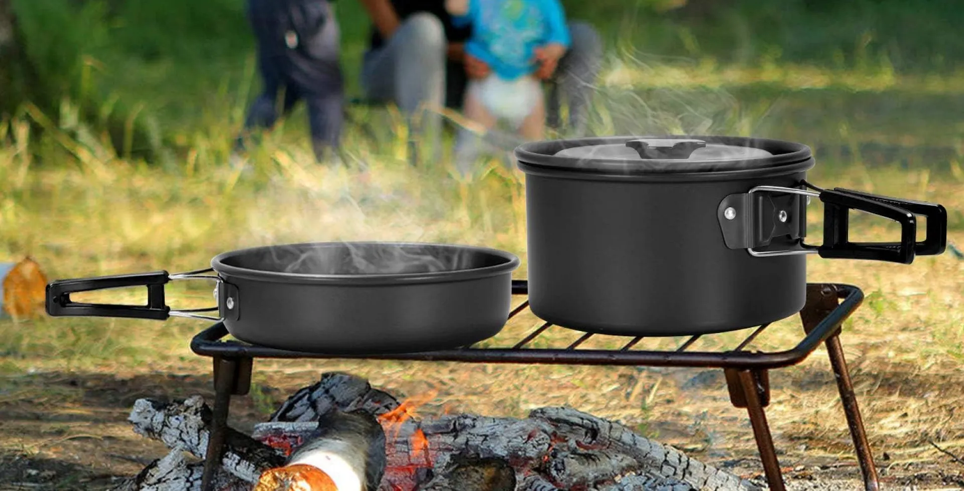 Odoland camping cookware set with pot and pan heating over open fire on a grill rack, steaming hot in a forest campsite with family in the background.