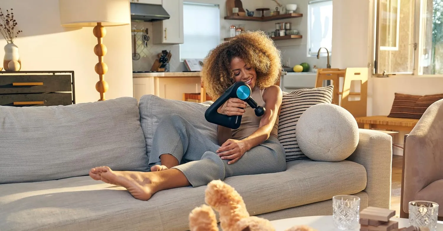 Woman using Theragun PRO massage gun on her arm while relaxing on a sofa in a sunlit modern living room – ideal for at-home muscle recovery and deep tissue therapy.